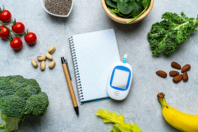 Healthy fruits and vegetables surrounding a blood sugar monitor, pen, and notepad