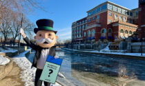 The Monopoly Man standing on a on the Naperville Riverwalk while holding a Naperville Riverwalk game piece.