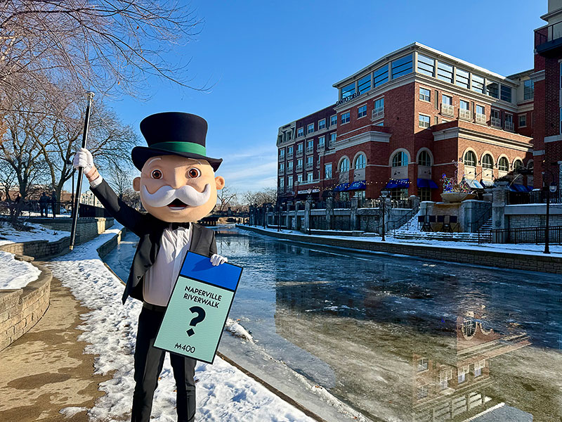 The Monopoly Man standing on a on the Naperville Riverwalk while holding a Naperville Riverwalk game piece.