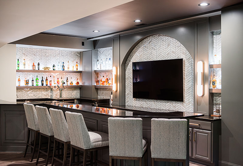 The next challenge was how to update the 2000s-era bar without busting the budget. “The cherry wood cabinets were very high-end, they were just outdated,” Karlye says. Silver suggested they keep the neutral black granite countertops and give the cabinetry a face-lift with new paint and hardware. “They had a ton of bottles, so we wanted to get them off the counter to declutter the space and use that height that they had here,” says Silver, who designed custom backlit shelving to display their bottles like artwork. “They also had a few different ceiling heights and soffits in this space, and we wanted to make it feel like it was all a part of the cabinetry.”
