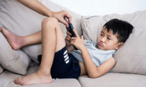 A parent's hand taking a tablet away from a pouting boy laying on a couch