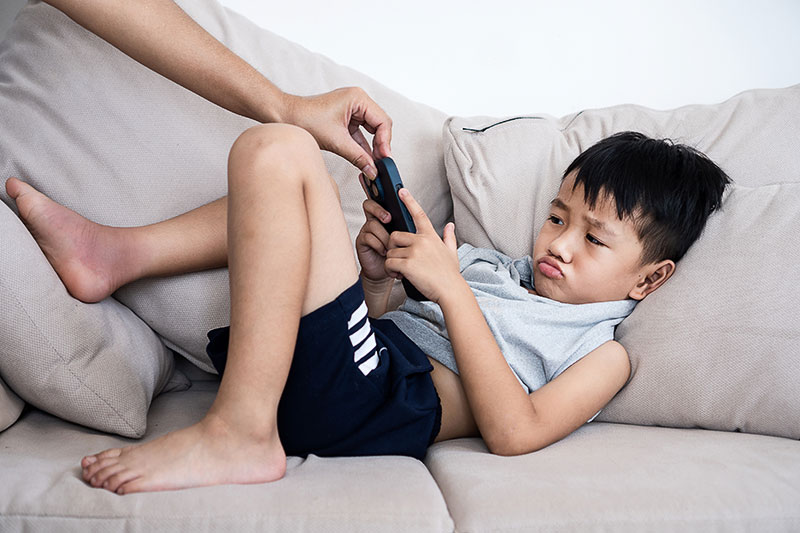 A parent's hand taking a tablet away from a pouting boy laying on a couch